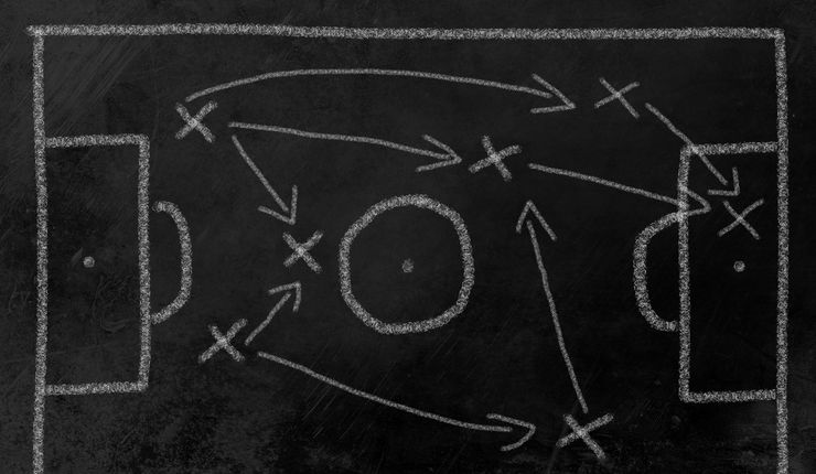Das Bild zeigt eine schwarze Tafel, auf der mit Kreide ein Fußballfeld mit Toren und Mittellinie schematisch dargestellt ist. Mit Kreuzen sind die Positionen einzelne Punkte markiert, die die Standorte von Spielern symbolisieren. Pfeile zwischen den Kreuzen markieren die Bewegungsrichtung der Spieler.