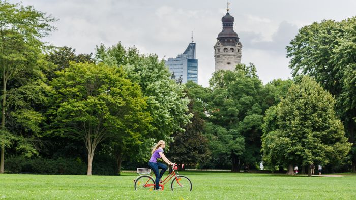 Junge Frau fährt mit dem Fahrrad durch den Park. Im Hintergrund ist das Neue Rathaus in Leipzig zusehen.