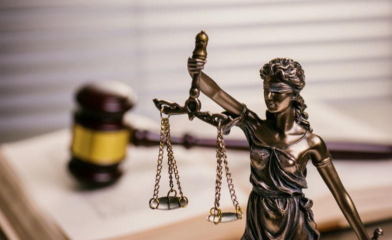 Bild zeigt eine Figur der Justitia, vor einem offenen Gesetzbuch, auf dem ein Richterhammer liegt