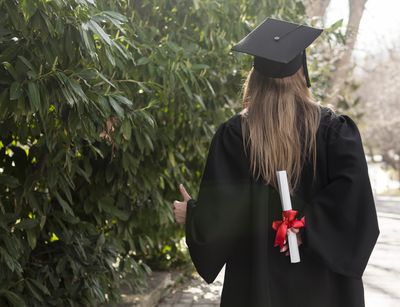 zeigt Frau in Alumni-Outfit von hinten mit einer Urkunde in der rechten Hand und einem Daumen nach oben der linken Hand