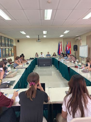 Etwa 20 Personen sitzen an einem Tisch. Sie führen ein Planspiel durch. Im Hintergrund hängen die Porträts verschiedener männlicher Personen. 