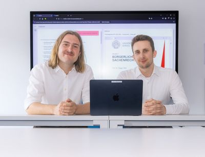 Zwei junge Männer sitzen an einem Tisch vor einem Großbildschirm, auf dem eine Internetseite zu sehen ist. 
