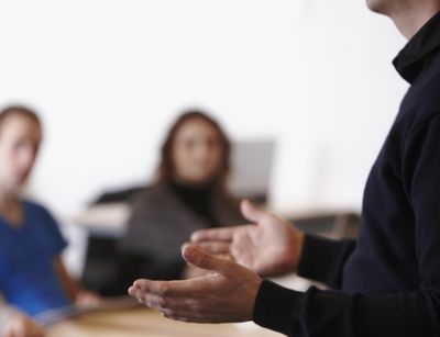 Auf dem Farbfoto ist eine Seminarsituation zu sehen. Im Vordergrund sind scharf die Hände eines Dozenten zu erkennen, im Bildhintergrund unscharf drei Studierende.