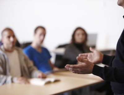 Auf dem Farbfoto ist eine Seminarsituation zu sehen. Im Vordergrund sind scharf die Hände eines Dozenten zu erkennen, im Bildhintergrund unscharf drei Studierende.