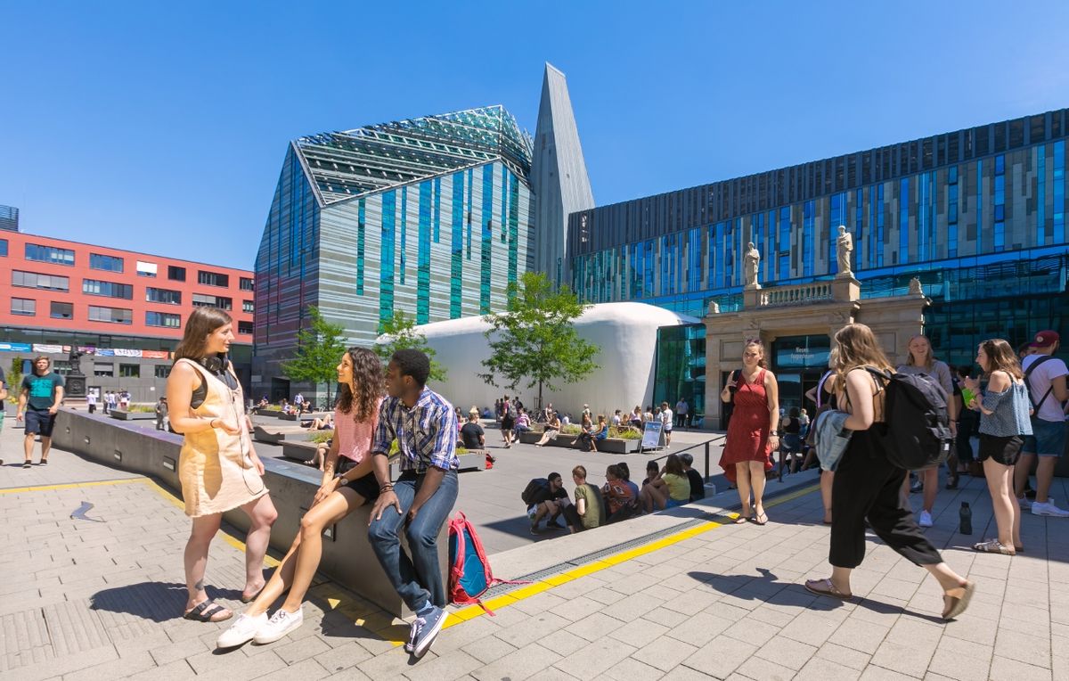 zur Vergrößerungsansicht des Bildes: Farbfotografie vom belebten Innenhof des Campus Augustusplatz in sommerlicher Atmosphäre. Im Fokus des Bildes steht eine Gruppe von drei jungen Menschen, die sich unterhalten. Zwei von Ihnen sitzen auf einer halbhohen Mauer. Eine weitere junge Frau steht ihnen zugewandt und spricht mit ihnen. Im Hintergrund sieht man mehrere kleine Gruppen von Menschen, die auf dem Campus verweilen oder über den Campus laufen.