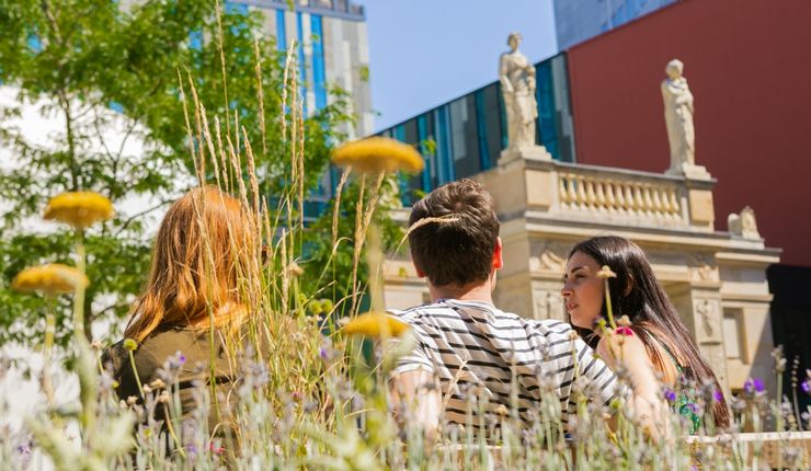 Drei Studierende sitzen auf dem Campus und unterhalten sich, im Hintergrund das Augusteum der Universität Leipzig.