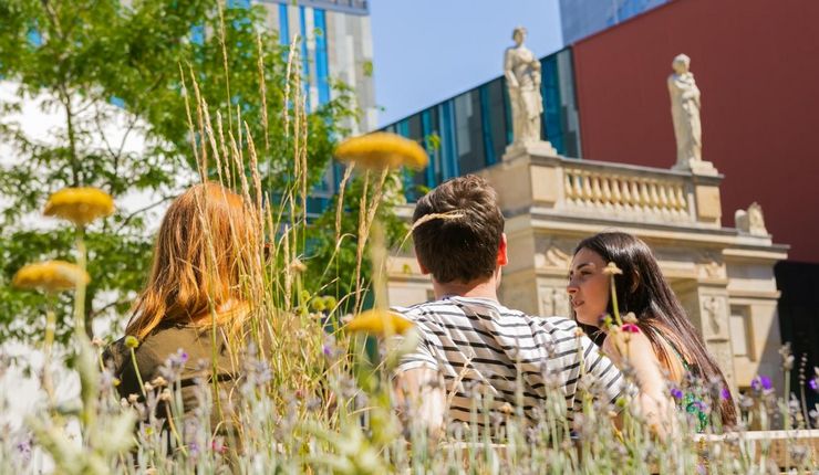 Drei Studierende sitzen auf dem Campus und unterhalten sich, im Hintergrund das Augusteum der Universität Leipzig.