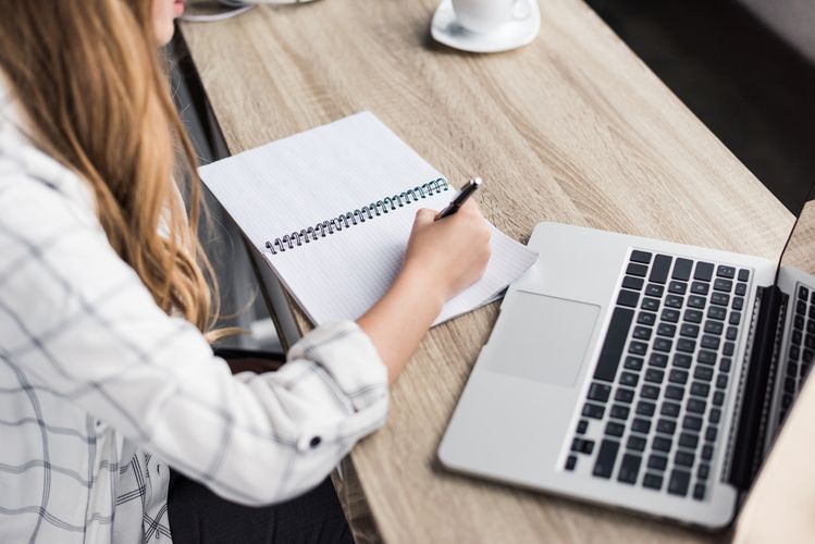 Frau an einem Schreibtisch macht Notizen in einem aufgeschlagenem Notizblock und Laptop