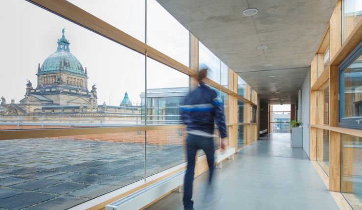 Foto: ein Mann ist auf den Gängen eines Gebäudes unterwegs. Durch die Glasfasade sieht man im Hintergrund das Bundesverwaltungsgericht.