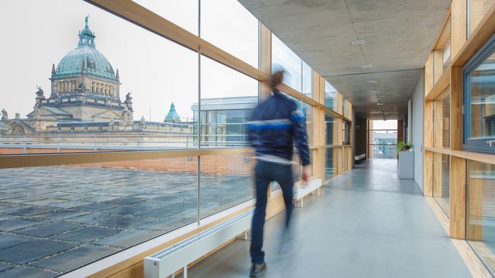 Foto: ein Mann ist auf den Gängen eines Gebäudes unterwegs. Durch die Glasfasade sieht man im Hintergrund das Bundesverwaltungsgericht.