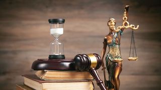 Auf dem Bild ist ein Stapel mit Büchern zu sehen. Auf diesem liegt eine Unterlage für einen Richterhammer. Auf dieser Unterlage steht eine umgedrehte Sanduhr. Gegen die Büchstapel gegen gelehnt ist ein Richterhammer. Neben diesem steht eine Statue, welche Justizia darstellen soll und folglich eine Wage in der linken Hand hoch hält und in der rechten Hand ein Schwert hält.