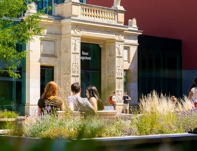 Farbfoto: Aufnahme des historischen Schickeltors im Leibnizforum am Campus Augustusolatz. Im Hintergrund ist der historische Torbogen zu sehen, davor sitzen mehrere Studierende auf Bänken, eingerahmt von einem bunt blühenden Blumenkasten und einem grünen Baum am linken Bildrand.