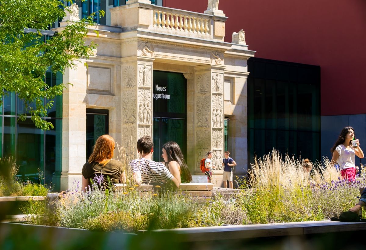 zur Vergrößerungsansicht des Bildes: Farbfoto: Aufnahme des historischen Schickeltors im Leibnizforum am Campus Augustusolatz. Im Hintergrund ist der historische Torbogen zu sehen, davor sitzen mehrere Studierende auf Bänken, eingerahmt von einem bunt blühenden Blumenkasten und einem grünen Baum am linken Bildrand.