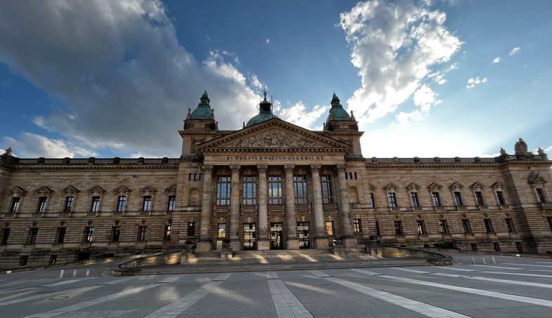 Bild des Einganges und Vorplatzes des Bundesverwaltungsgerichts in Leipzig mit blauem Himmel, einzelnen Wolken und dem Schein der Sonne, die hinter dem Gebäude steht.