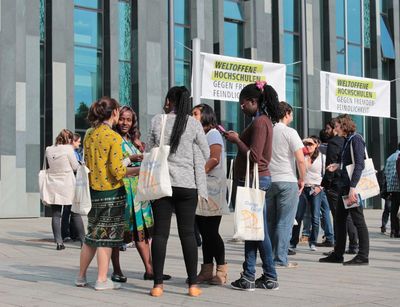 Zu sehen auf dem Foto ist eine internationale Menschengruppe auf dem Augustusplatz, im Hintergrund ist das Universitätsgebäude sichtbar. Die Personen scheinen im regen Austausch zu sein, im Hintergrund prangen Plakate mit der Aufschrift "Weltoffene Hochschulen gegen Fremdenfeindlichkeit".