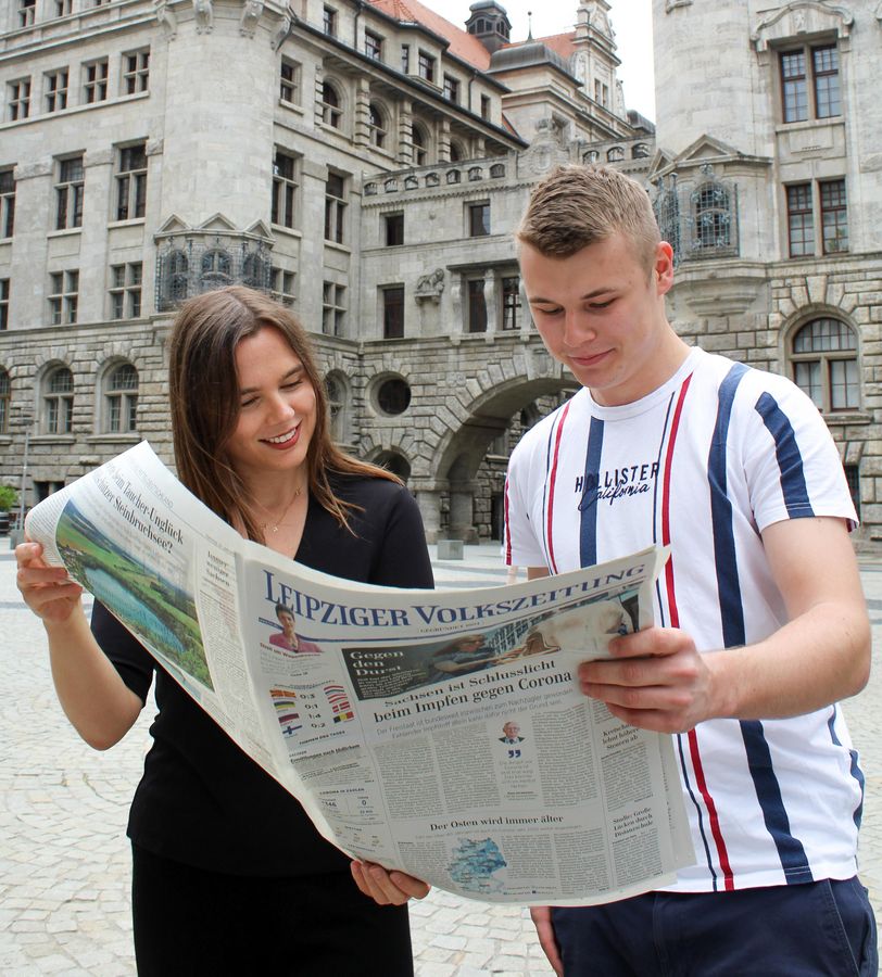 Laura Barth und Yannis Nehrig blicken auf eine Zeitung, im Hintergrund: neues Rathaus Leipzig