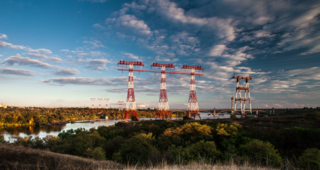 Landschaftsfotografie: Hoschpannungsleitungen in einer Landschaft bei Sonnenuntergang, im Hintergrund ist eine Flusslandschaft zu sehen, im Vordergrund stehen mehrere Hochspannungsmasten