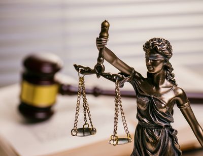 Bild zeigt eine Figur der Justitia, vor einem offenen Gesetzbuch, auf dem ein Richterhammer liegt