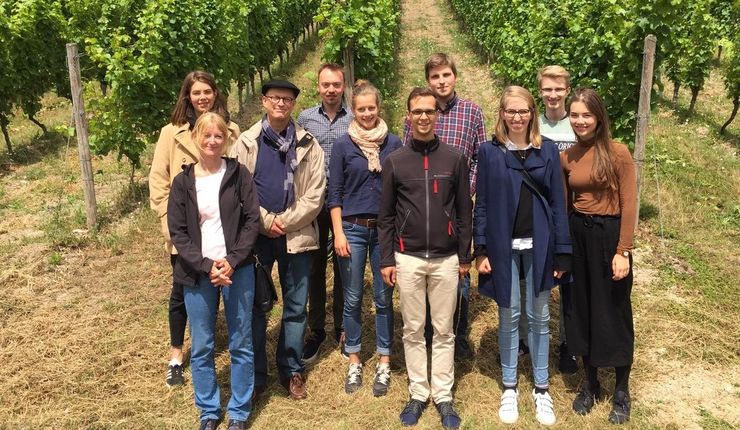 Eine Gruppe von Personen gemischten Alters und gemischten Geschlechts steht vor einem sonnigen Weinhang und lächelt in die Kamera.